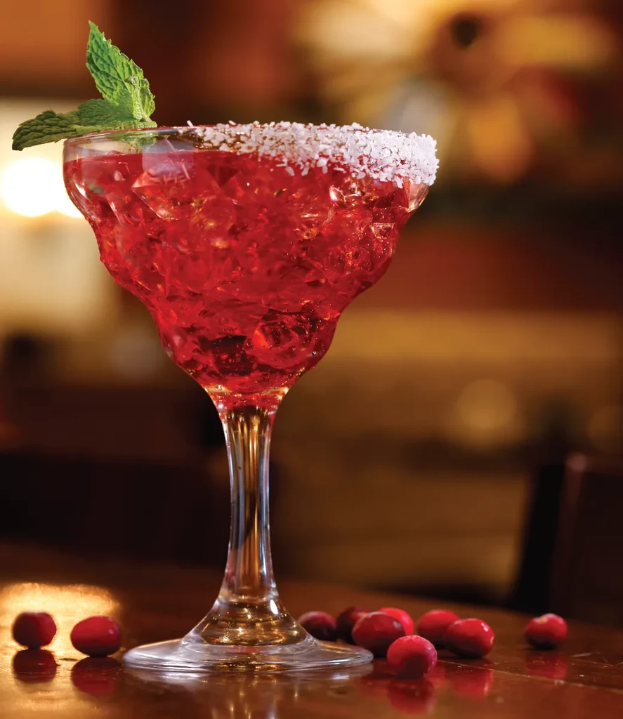 Red cocktail in margarita glass, sugared rim, mint, cranberries on wood.