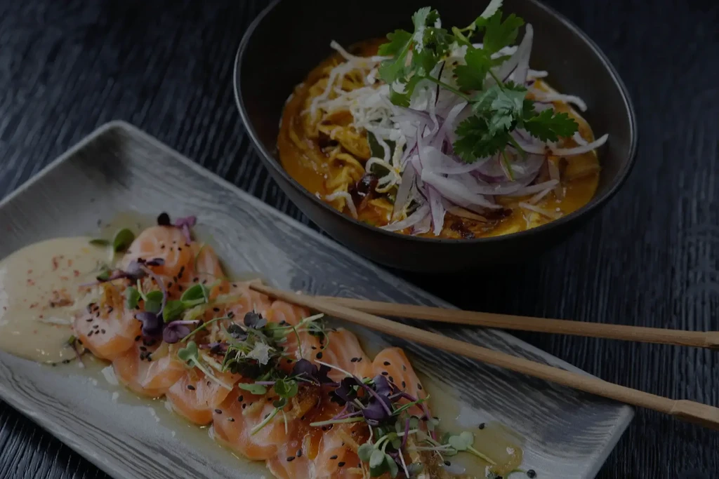 Salmon sashimi with chopsticks and a noodle bowl on dark surface.
