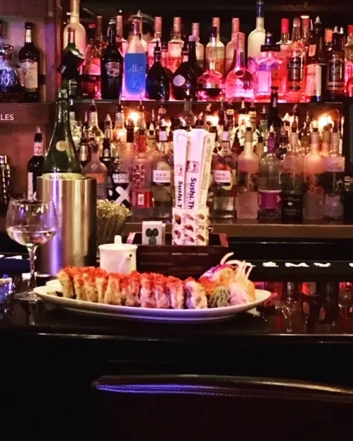 Sushi, wine, and tea at a dimly lit bar with colorful bottles.
