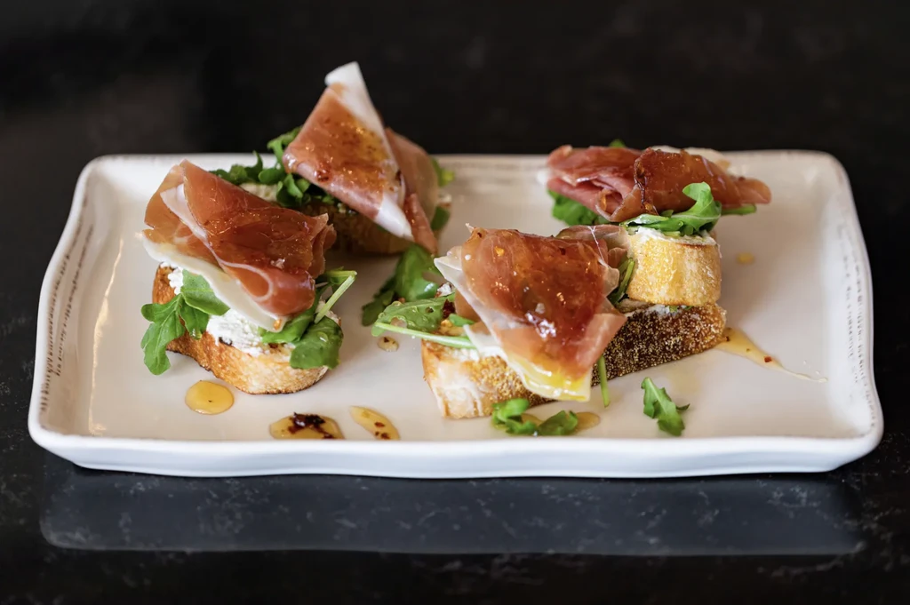 Five crostini with cheese, prosciutto, arugula, and amber drizzle on white platter.
