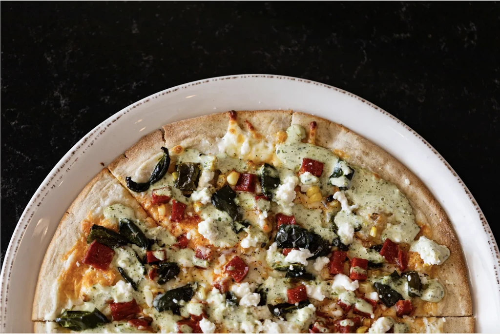 Overhead colorful pizza with green sauce, white cheese, peppers, and corn.