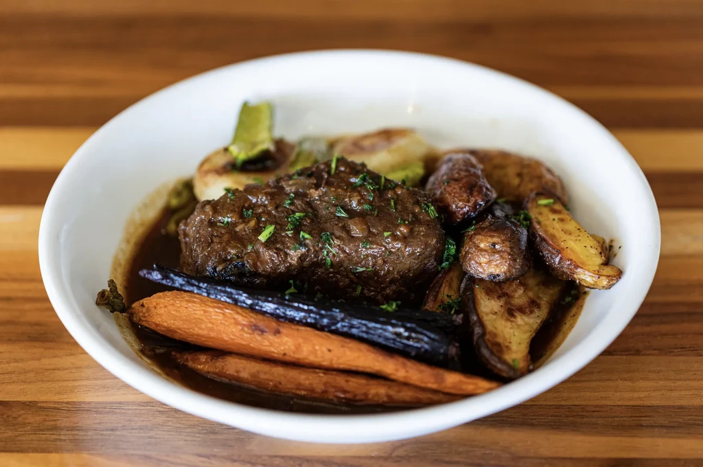 Braised meat, roasted carrots, potatoes, and herbs in a white bowl.