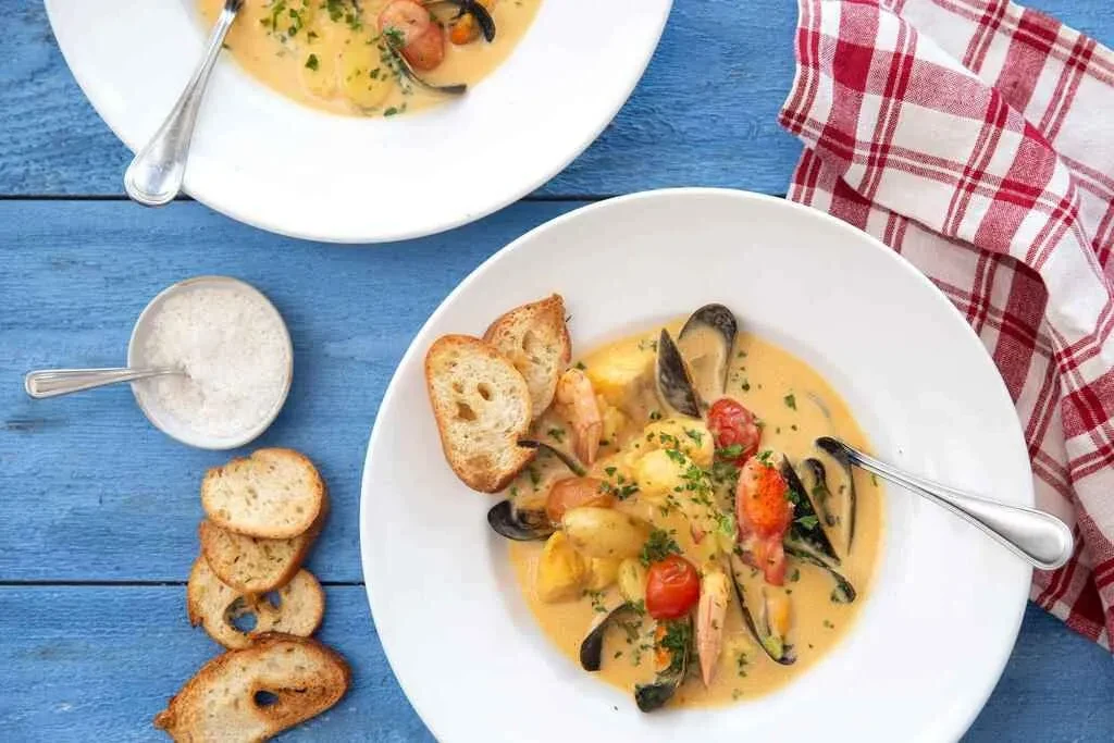 White bowl of creamy seafood stew with mussels, shrimp, baguette.