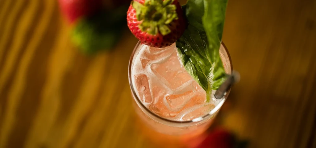 Overhead pink drink with strawberry, basil, ice, straw on wood.
