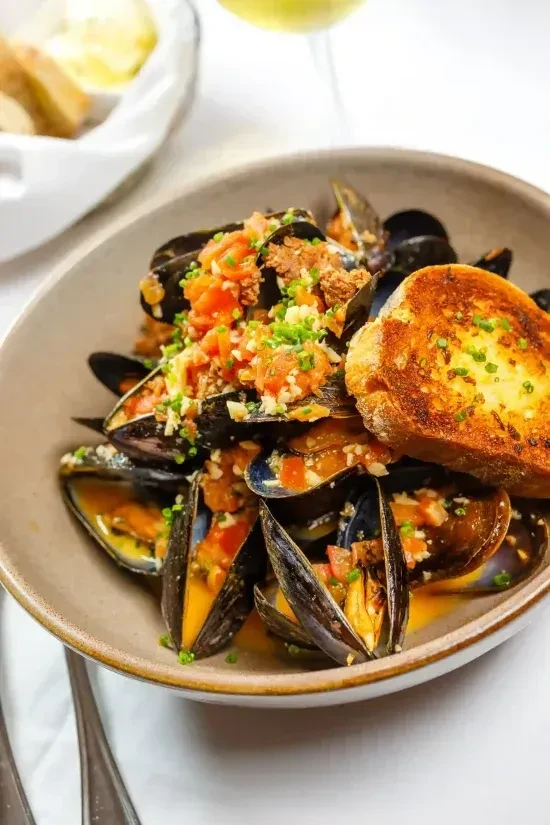 Mussels in yellow broth, garnished with tomatoes, chives, toasted bread.
