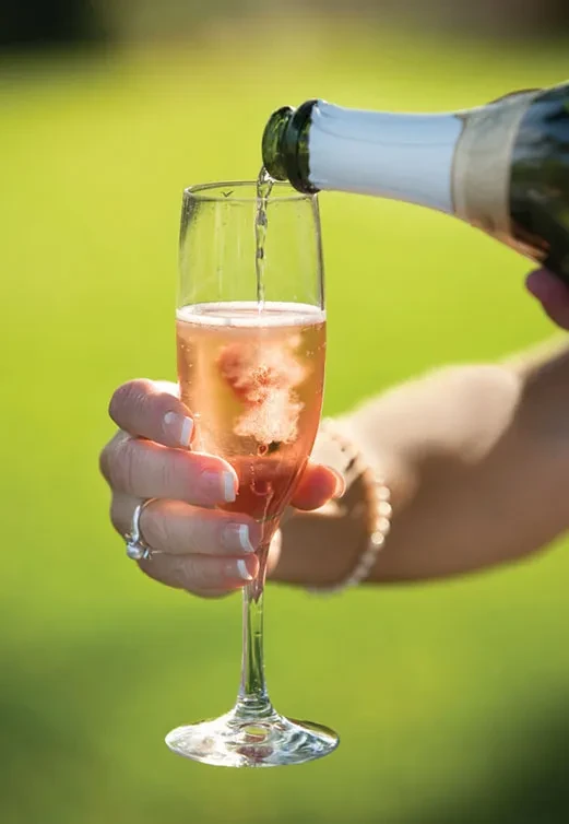 Rosé sparkling wine pours into flute, held by hand, with fruit garnish.