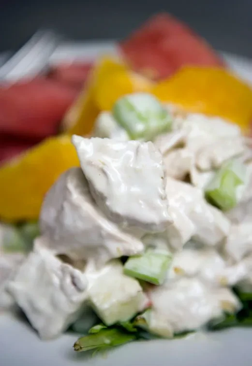 Close-up of creamy chicken salad with celery on plate, fruit background.
