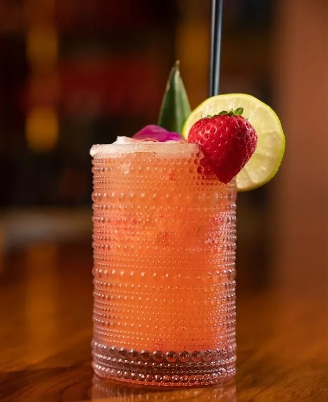 Orange-pink cocktail in patterned glass, garnished with strawberry, lime, leaf, flower.