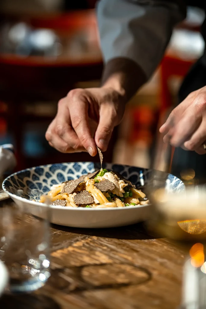 Close-up of hands garnishing creamy pasta with black truffle slices.