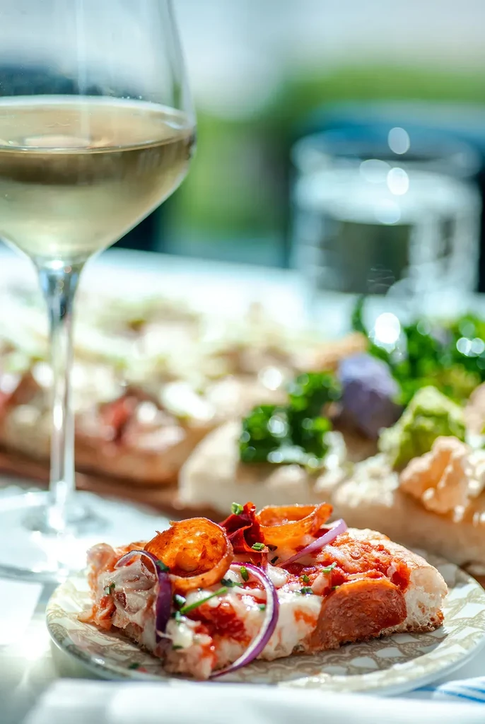 Close-up of a pepperoni pizza slice, white wine, and blurred food spread.
