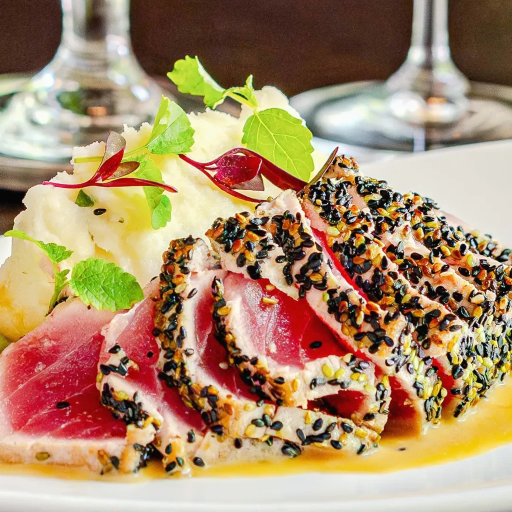 Plated seared tuna with sesame crust, mashed potatoes, sauce, and microgreens.