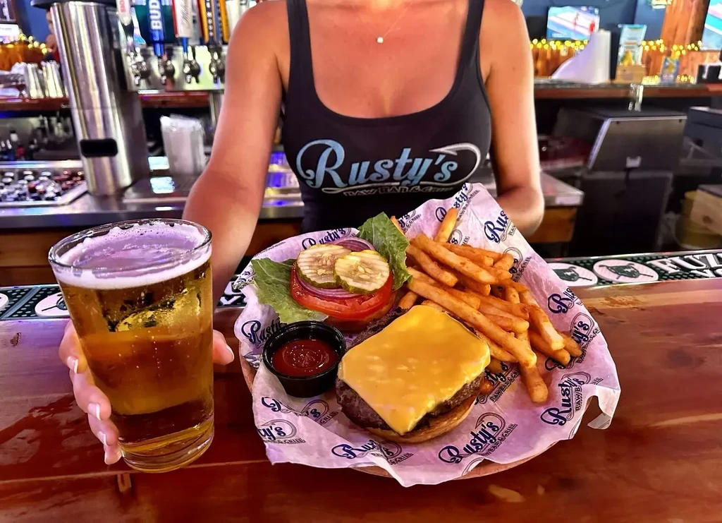 Person holds cheeseburger, fries, and beer at Rusty's Raw Bar.