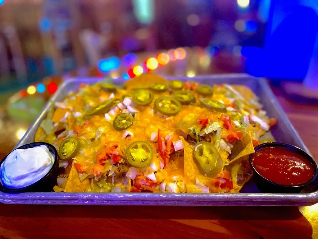 Nachos in metal tray with cheese, jalapeños, tomatoes, lettuce, and dips.