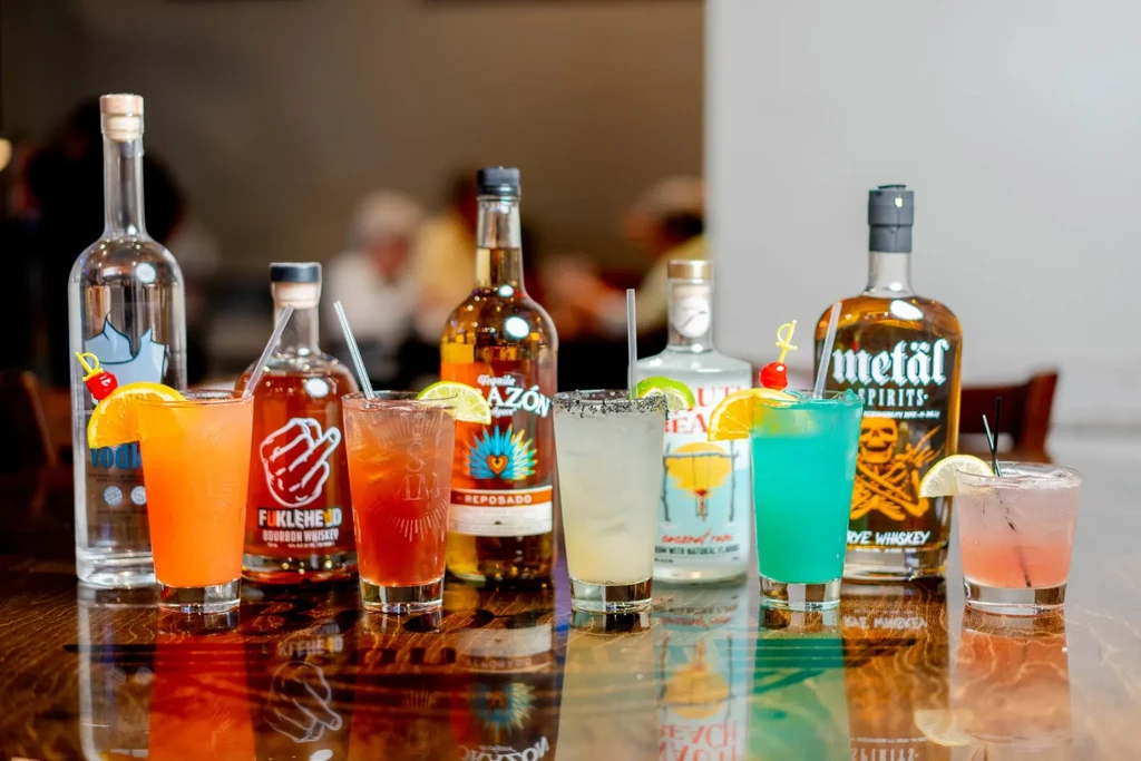 Seven garnished cocktails and five liquor bottles on a reflective wooden table.
