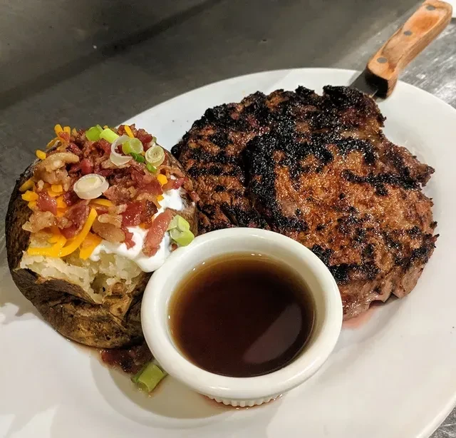 White plate with grilled steak, loaded baked potato, and dipping sauce.