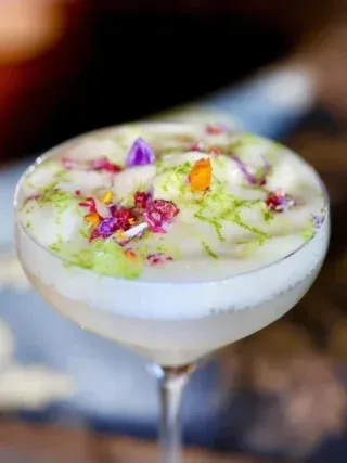 Frothy cocktail in coupe glass, garnished with colorful zest and flower petals.