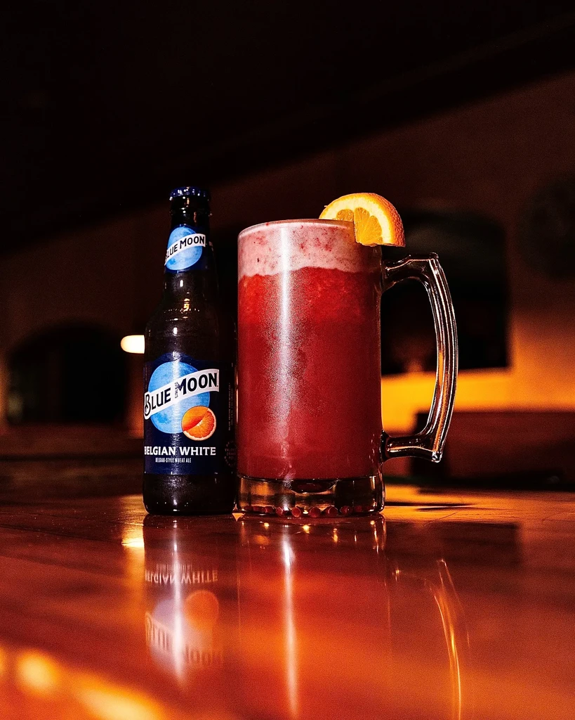 Blue Moon bottle and frosty purple drink with orange on a bar.