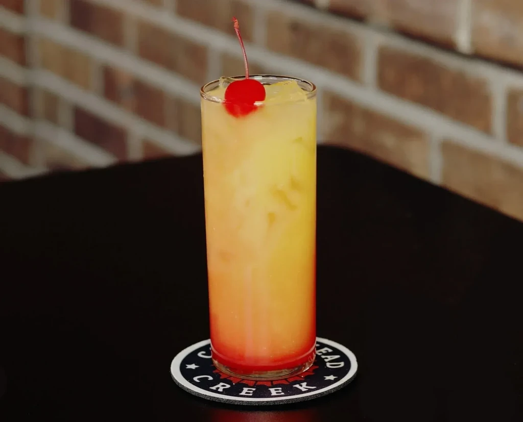 Layered red-orange-yellow cocktail with cherry, ice, and Creek Head coaster.