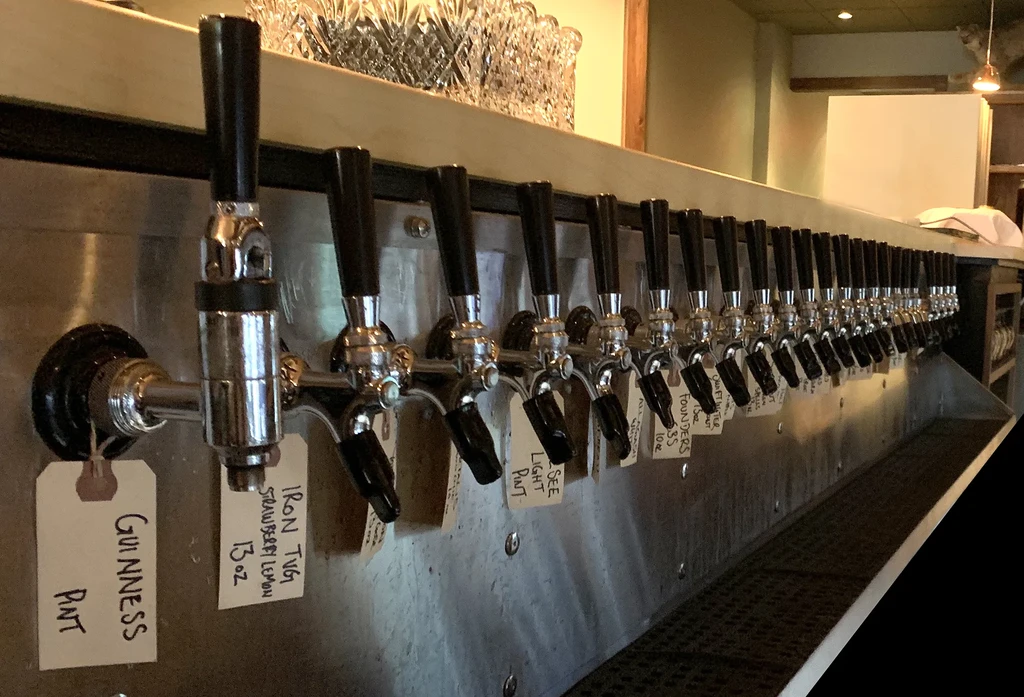 Long row of chrome beer taps with handwritten labels on a bar.