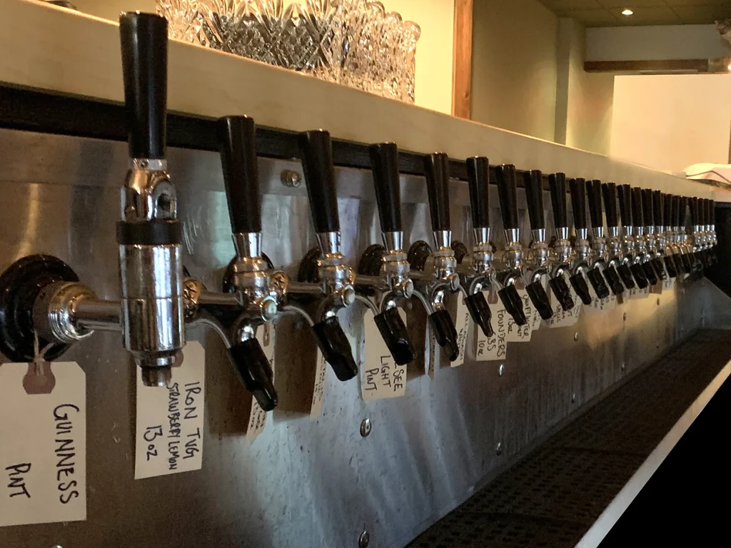 Numerous chrome beer taps with handwritten tags on a stainless bar.