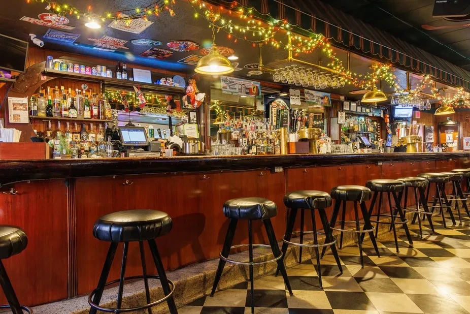 Well-lit empty bar with wooden counter, stools, liquor bottles, checkered floor.