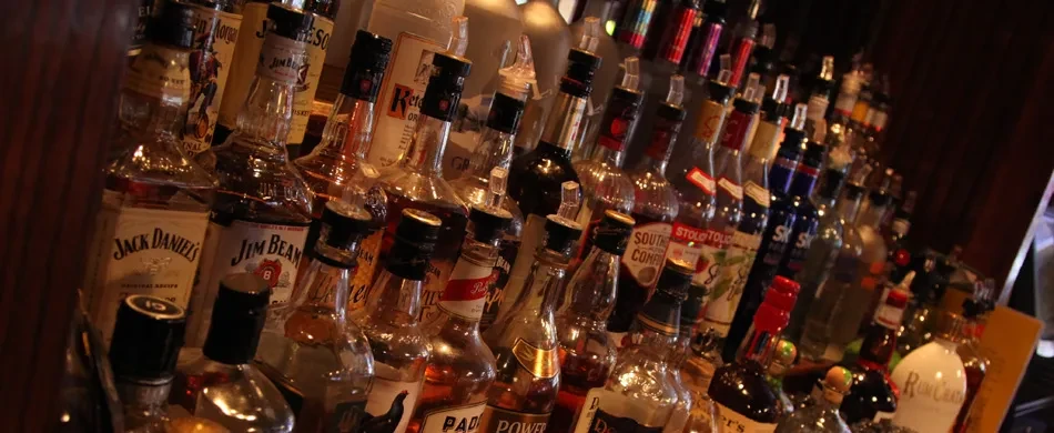 Many whiskey and spirits bottles with pour spouts on a dim bar.