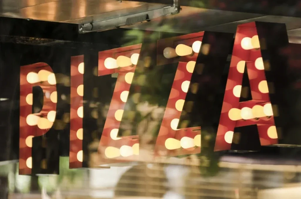 Red "PIZZA" marquee sign with glowing bulbs, viewed through a window.