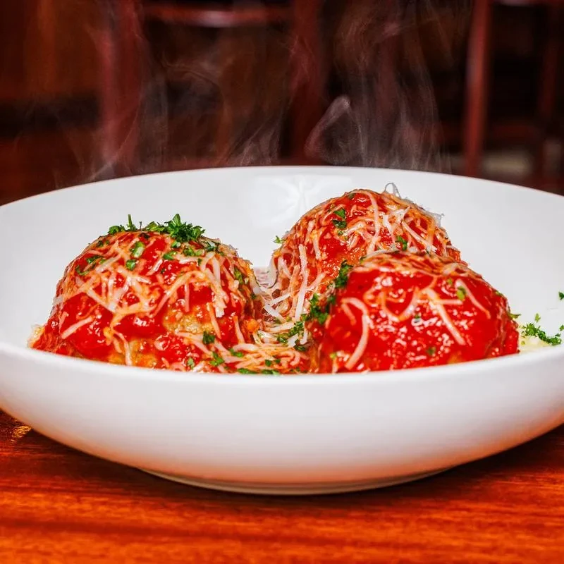 Three steaming meatballs with tomato sauce, cheese, and herbs in a white bowl.