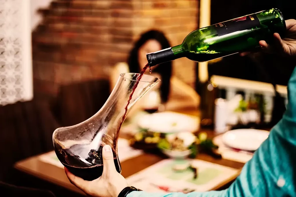 Close-up of hands pouring red wine into a glass decanter.