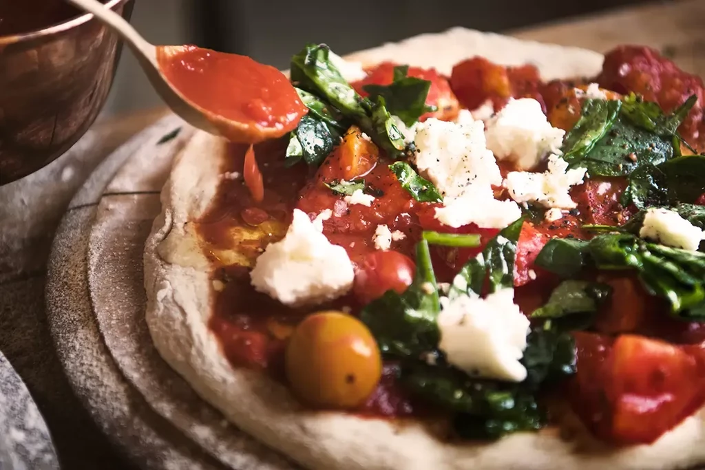 Fresh uncooked pizza with sauce, tomatoes, spinach, cheese, spoon dripping.