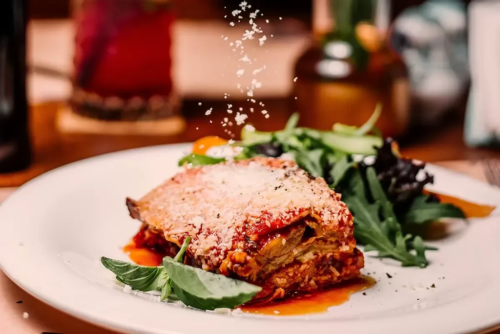Layered lasagna with grated cheese falling, fresh basil, and a side salad.