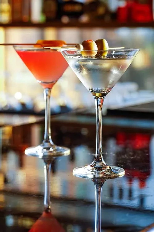 Two cold cocktails in martini glasses: clear with olives, pink with orange.