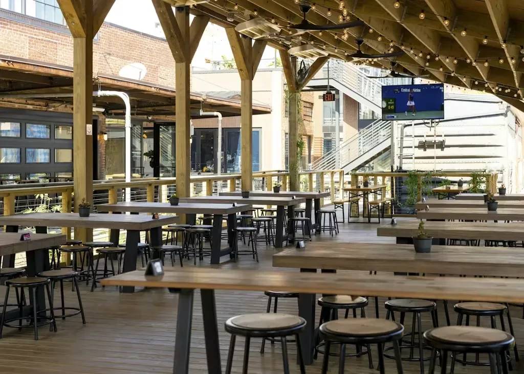 Outdoor patio with empty high-top tables, string lights, and tennis on TV.