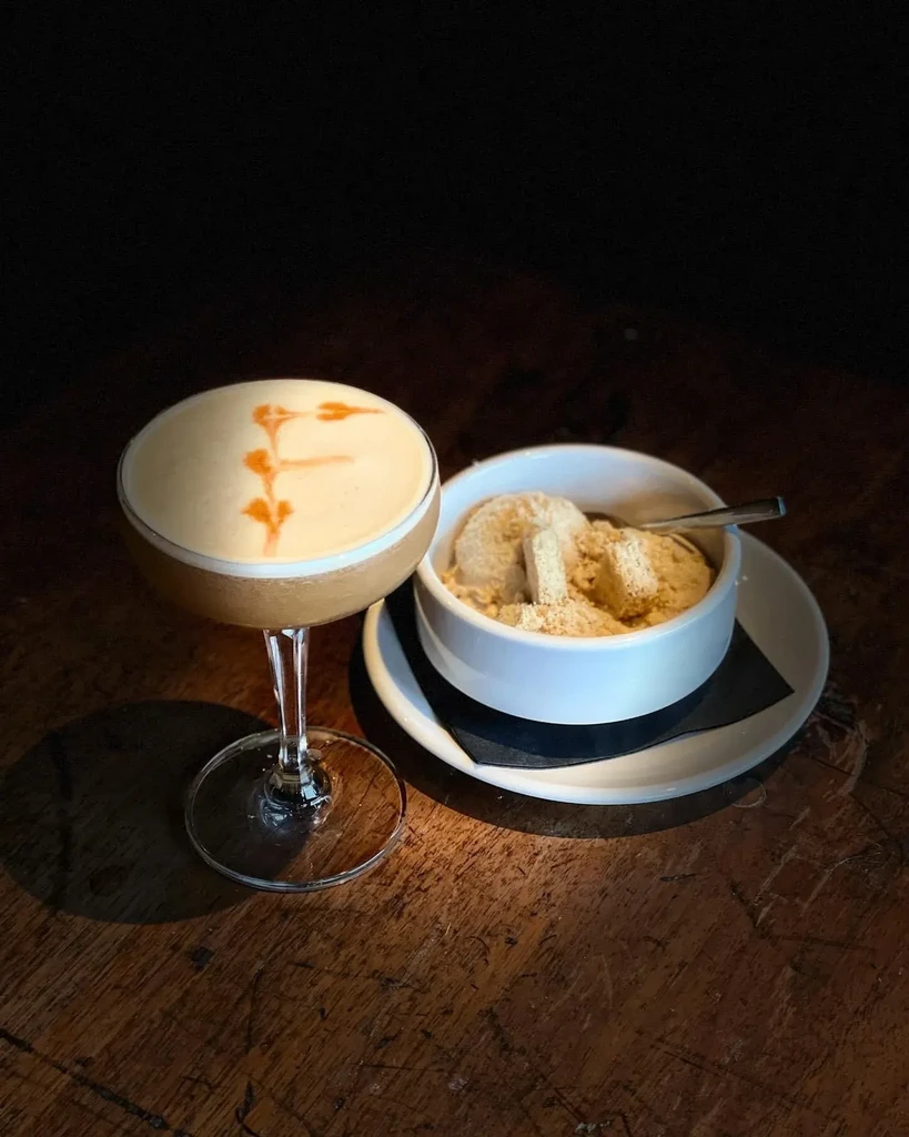 Foamy cocktail with 'F' and crumb-topped dessert on dark wood.