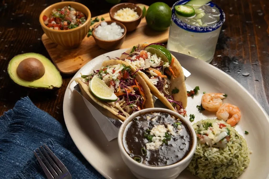 Two tacos, beans, rice, and shrimp served on a wooden table.