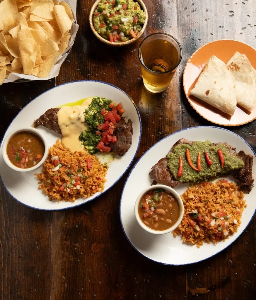 Two plates of Mexican cuisine, chips, guacamole, and a drink.