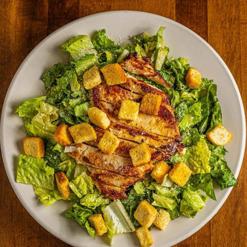 Caesar salad with grilled chicken, croutons, and parmesan on wooden table.