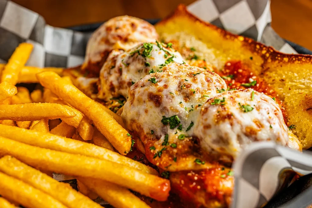 Close-up of meatballs with melted cheese, red sauce, fries, and garlic bread.