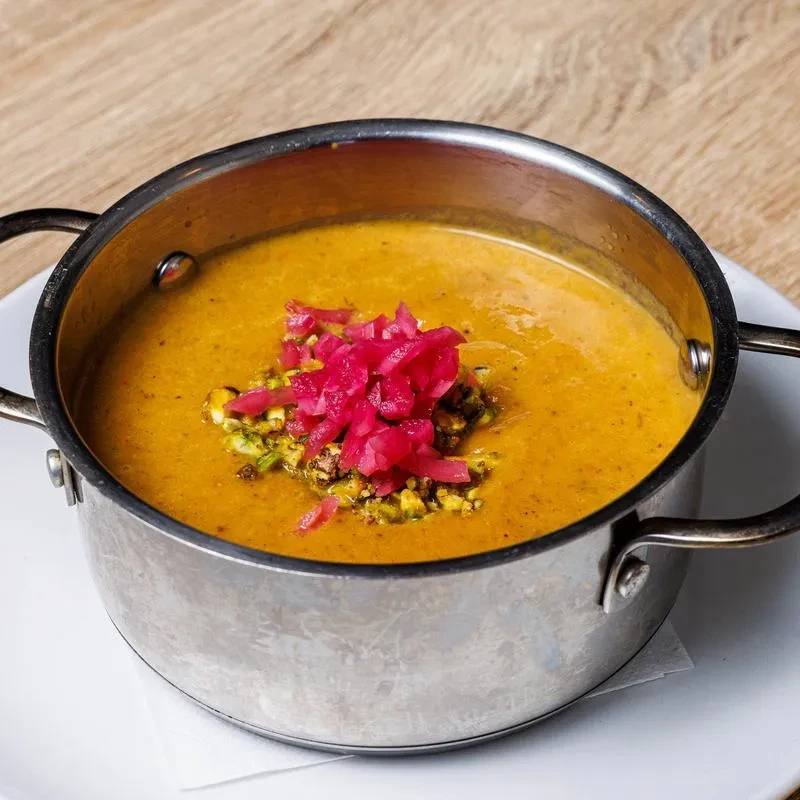 Creamy golden-orange soup in steel pot with green and pink garnishes.