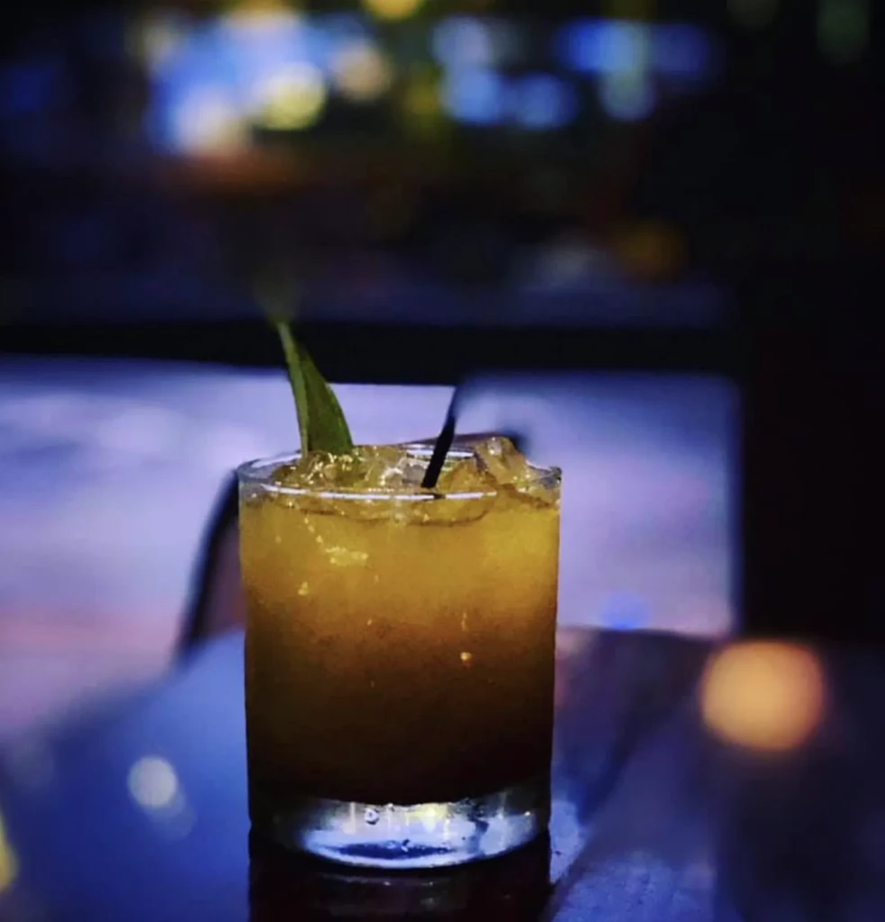 Amber cocktail in rocks glass with ice, leaf, and black straw.