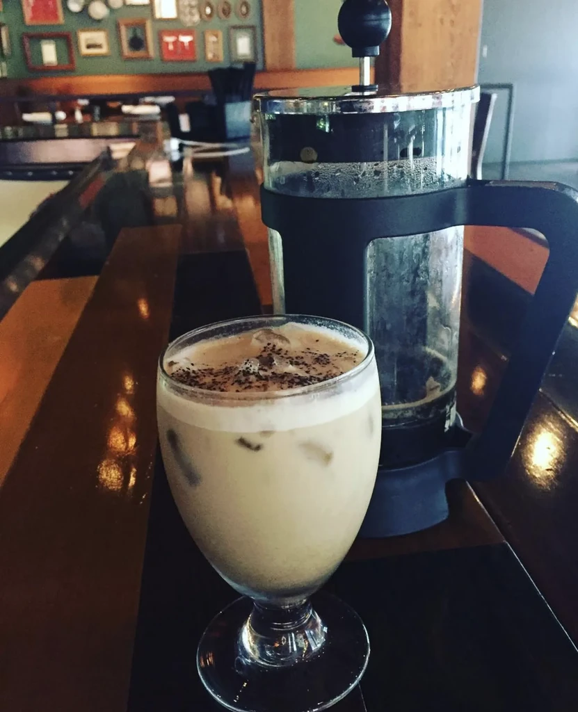 Frothy beverage in stemmed glass, French press behind in cafe.
