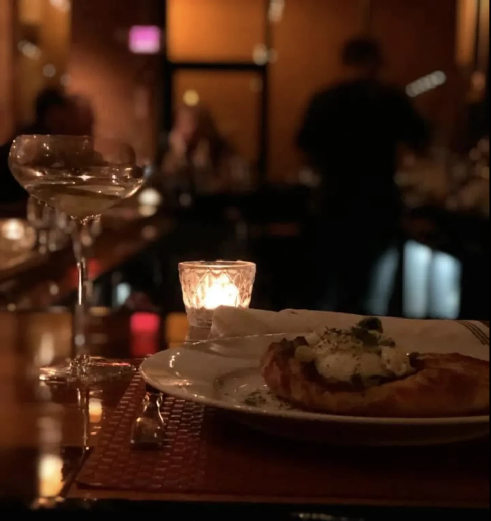 Warmly lit restaurant table with a cocktail, candle, and savory pastry.