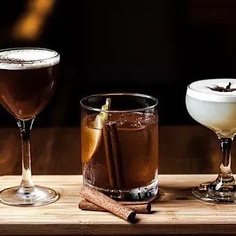 Three cocktails on wood: a dark foam, dark cinnamon, and creamy anise.