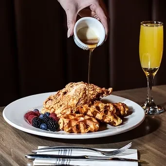Hand pouring syrup over fried chicken and waffles with berries and mimosa.