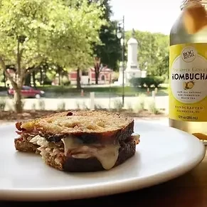 Close-up of Reuben-style sandwich, kombucha bottle, and outdoor window view.