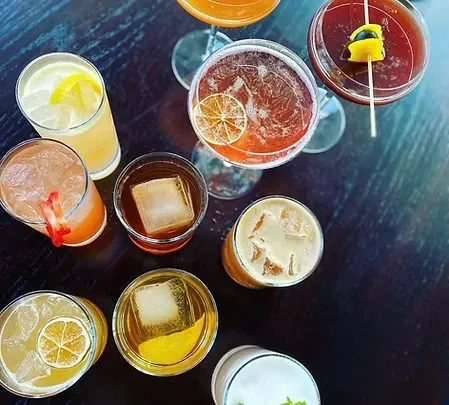 Overhead shot of nine colorful, garnished cocktails on a dark tabletop.