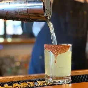 Cocktail shaker pouring yellow liquid into a chili-salt-rimmed glass.