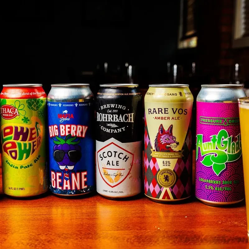 Five distinct beer cans and a partial glass on a wooden bar.