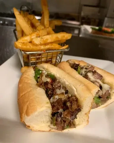 Two cheesesteak-style sandwiches, one bitten, with golden fries.