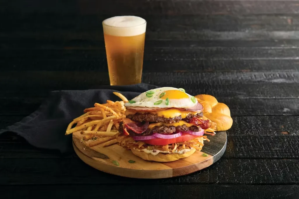 Stacked burger with egg, bacon, fries, beer on wooden board.
